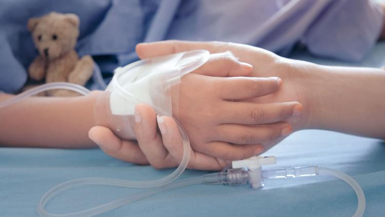 Close de uma das mãos de uma criança internada com linfohistiocitose hemofagocítica sendo segurada pela de um adulto